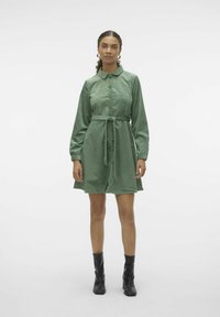 Robe chemise en velours côtelé vert avec un col, des boutons sur le devant, des manches longues et une ceinture assortie. Assortis avec des bottes à talons noires.