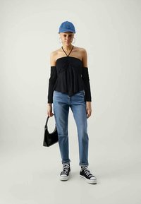 Zwarte off-the-shoulder top met lange mouwen, blauwe denim jeans, zwarte sneakers en een blauwe pet. Houdt een kleine zwarte handtas vast.