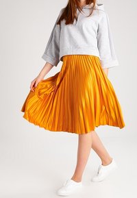 Mujer vestida con un crop hoodie gris y una falda plisada naranja brillante, de pie sosteniendo la falda con zapatos slip-on blancos.