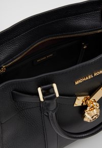 Borsa a mano in pelle nera con superficie texturizzata, hardware dorato, un lucchetto decorativo e una tasca con zip interna. Etichetta del marchio visibile all'interno.