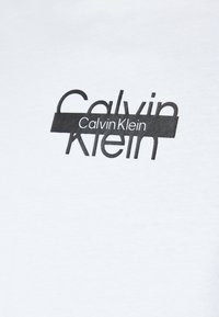 Hvit bomull T-skjorte med et svart grafisk trykk som har teksten "Calvin Klein" i varierende skrifttyper og størrelser.