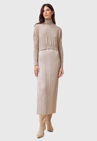 Maglione corto color beige con collo a imbuto abbinato a una gonna beige plissettata, caratterizzata da una texture liscia. Il modello indossa stivaletti alla caviglia beige coordinati.