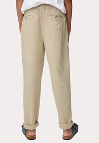 Pantalons en lin beige avec une taille élastique, dotés de deux poches arrière et de poignets retroussés. Associés à des chaussures bleues à enfiler.