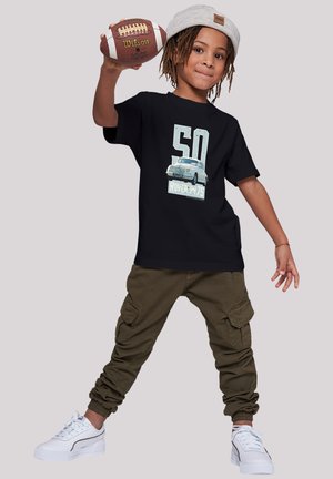 Black cotton t-shirt with a car graphic, cargo pants in olive green, and white sneakers. The child holds a brown football.