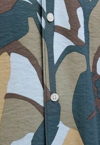 Gros plan d’un vêtement boutonné avec un motif abstrait multicolore dans des tons bleus, bruns et beiges sur tissu texturé.