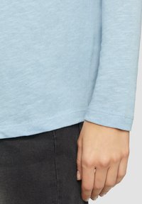 Hellblaues Langarmhemd aus weichem Stoff mit abgerundetem Saum und eingesetzten Ärmeln. Getragen mit dunkelgrauten Hosen.
