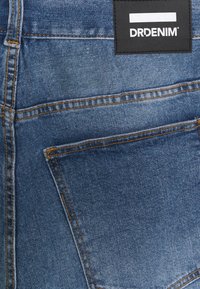 Poche arrière d'un jean en denim bleu avec des coutures orange et une étiquette rectangulaire noire portant l'inscription « DRDENIM » cousue au-dessus de la poche.