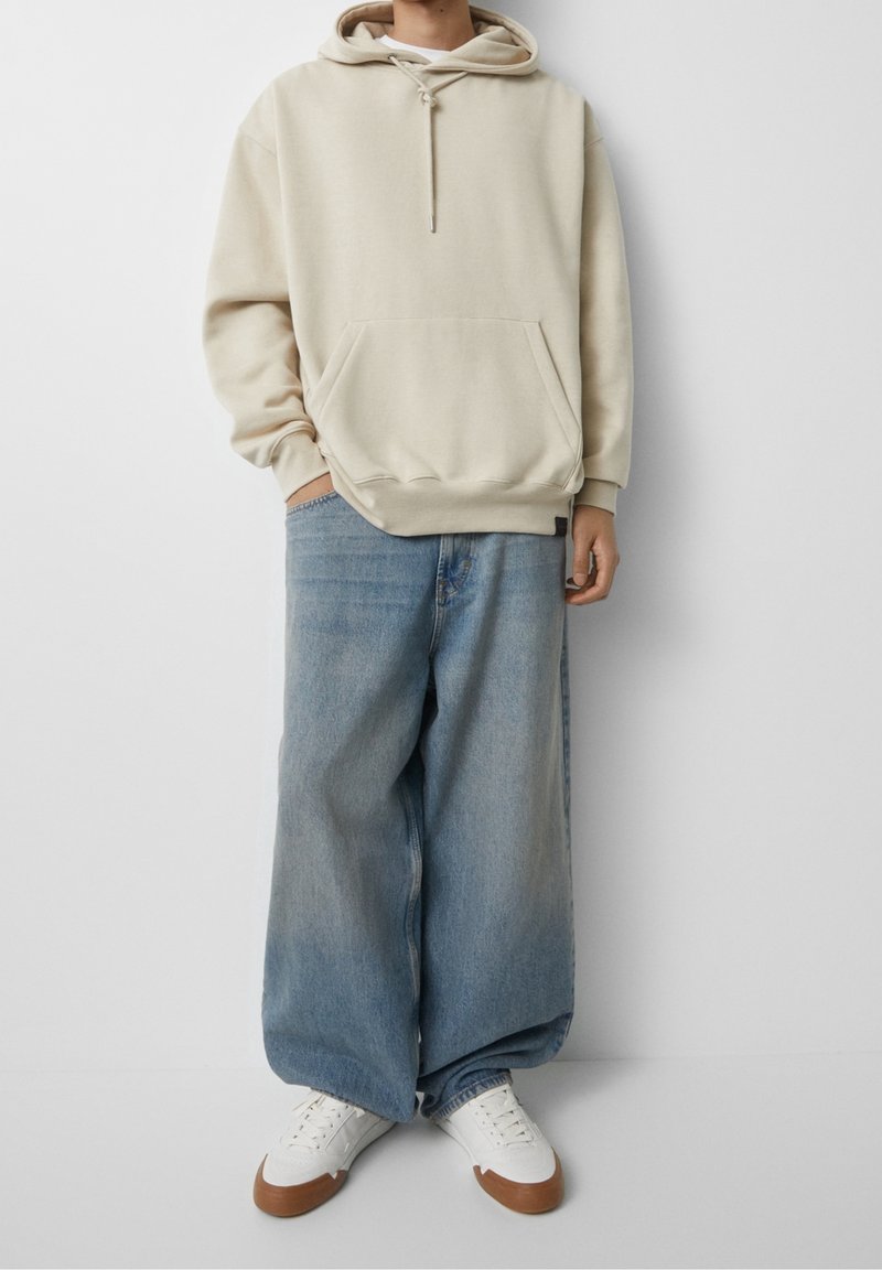 Personne portant un sweat à capuche beige, un jean large bleu clair et des baskets blanches avec des semelles marron, debout devant un fond uni.