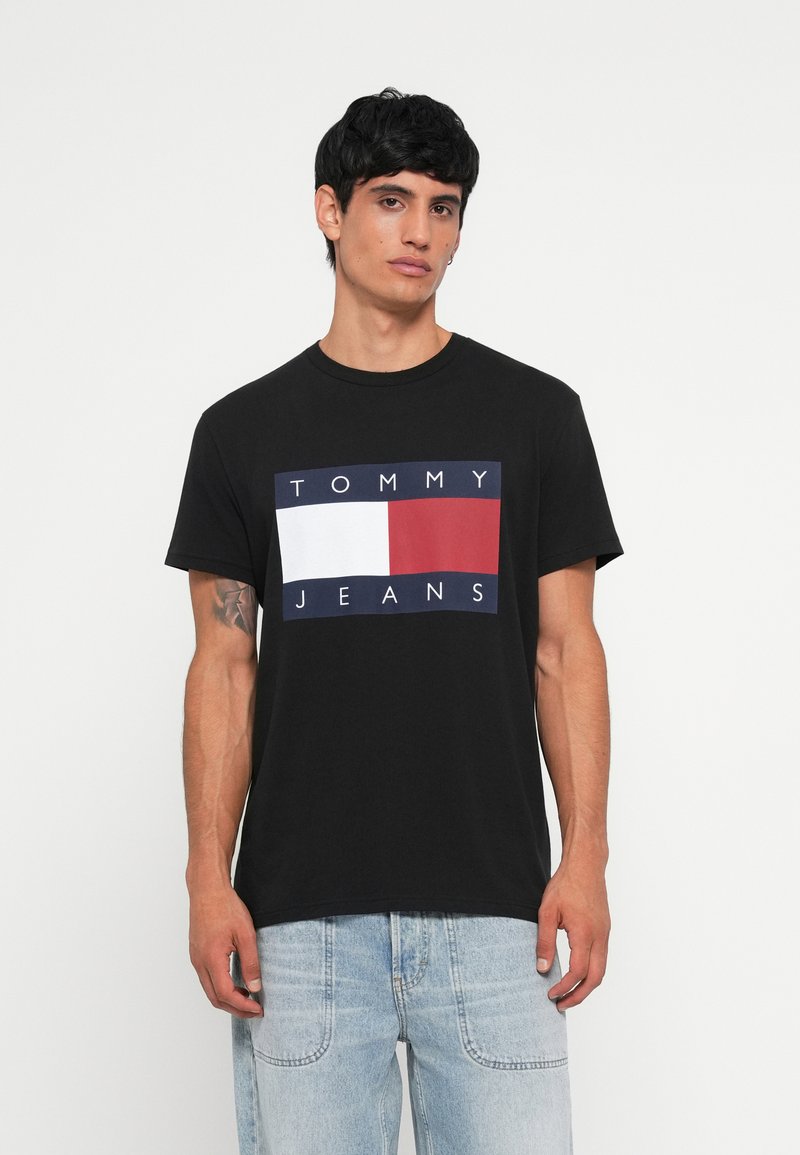 Must musta T-särk, millel on julge esikülg, kus on valge tekst "TOMMY" ja valge tekst "JEANS", mis on paigutatud punase ja valge ristküliku taustale.