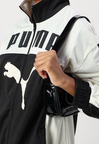 PUMA Windbreaker mit schwarz-weißen Farbkombinationen, oversized Logo, Reißverschluss und gerippten Bündchen. Hält eine glänzende schwarze Handtasche.