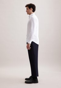 Witte, langelijf shirt met knoopaccenten op de manchetten, gecombineerd met marineblauwe broek en zwarte nette schoenen, weergegeven in zijaanzicht.