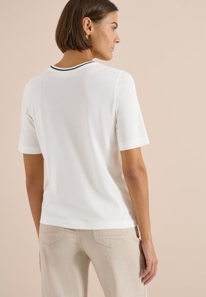 Femme aux cheveux courts et bruns portant une chemise blanche à manches courtes avec un bord noir au col et un pantalon beige, de profil.