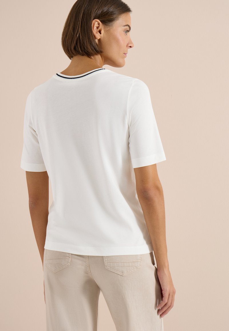 Femme aux cheveux courts et bruns portant une chemise blanche à manches courtes avec un bord noir au col et un pantalon beige, de profil.