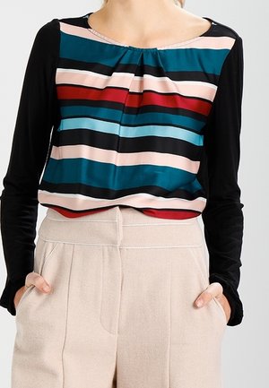 Kvinde iført langærmet bluse med farverige, vandrette striber og lyse beige bukser med høj talje, hænderne i lommerne.