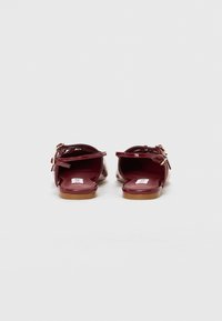 Sandales vernies bordeaux avec deux brides de cheville et une semelle plate. Présentent un accent de boucle et une texture lisse et brillante.