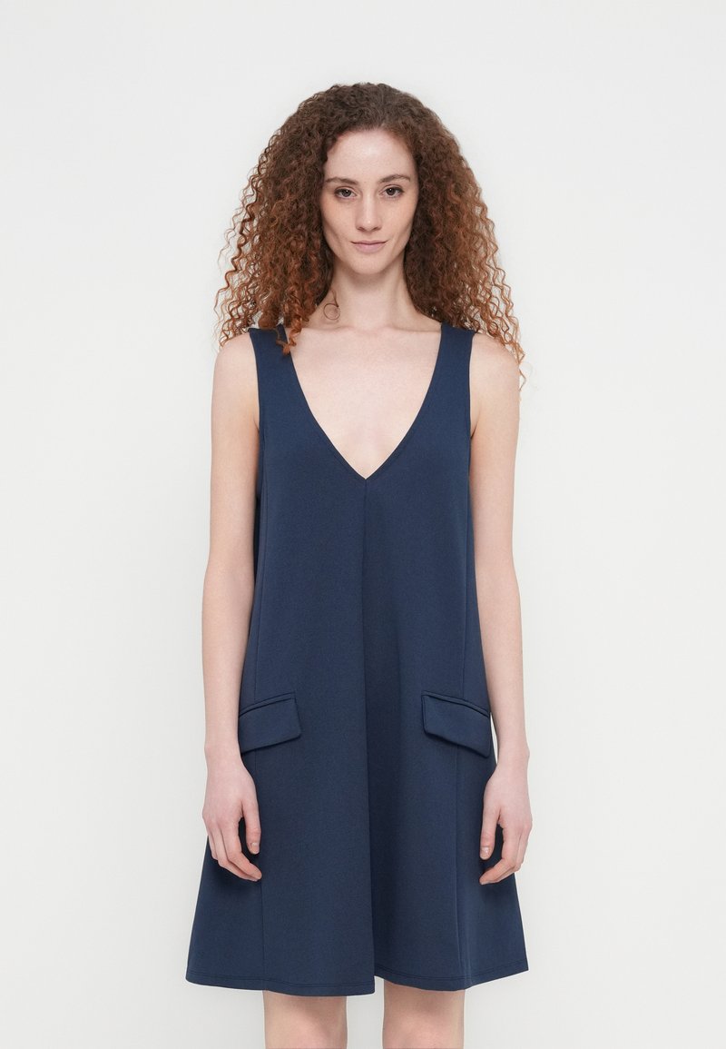 Γυναίκα με σγουρά κόκκινα μαλλιά, φοράει αμάνικο μπλε-ναυτικό φόρεμα με V-λαιμόκοψη και εμπρός τσέπες με καπάκι, στέκεται μπροστά σε λευκό φόντο.