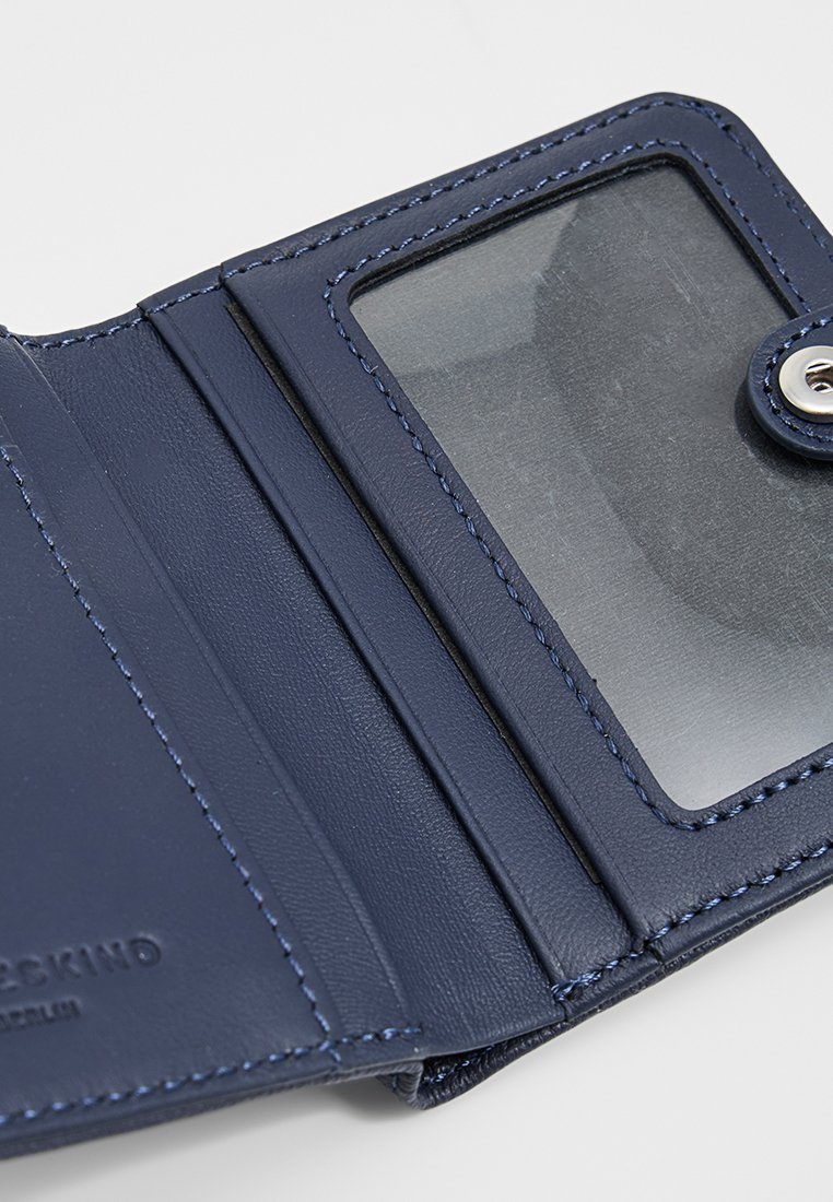 Granatowy skórzany portfel o gładkiej teksturze, z przezroczystą kieszonką na karty i detalami w postaci przeszyć. Zamyka się na guzik zatrzaskowy.
