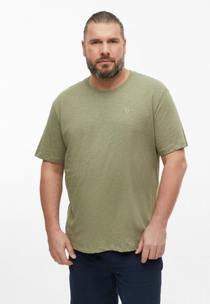 Man with beard and tied-back hair wears olive green short-sleeve shirt and dark pants, standing against a plain white background.
