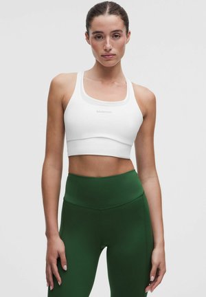 Kvinde iført hvid sports-bh og mørkegrønne leggings med høj talje, stående med neutralt udtryk mod en ensfarvet hvid baggrund.