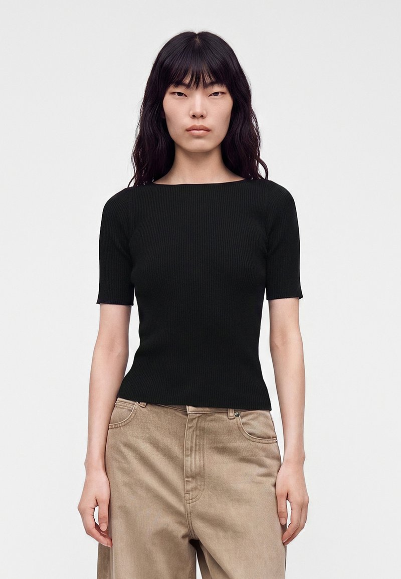 Femme aux longs cheveux noirs portant un haut noir ajusté à manches courtes et un pantalon beige ample, debout devant un fond clair uni.