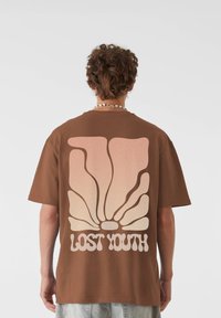 Barna, oversized t-shirt, amelynek hátulját egy nagy, pasztell színű, átmenetes kagyló grafika díszíti, alatta pedig a "LOST YOUTH" felirat található, vastag betűtípussal.