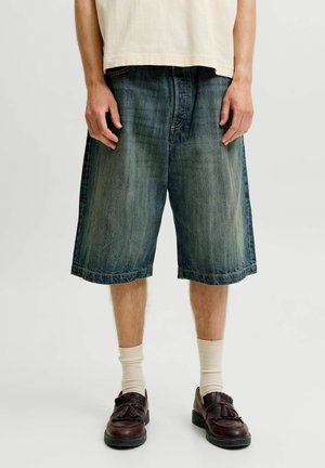 Personne portant un short en jean ample délavé, des chaussettes couleur crème, des mocassins à pampilles brun foncé et une chemise beige, debout devant un fond uni.