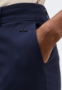 Χέρι στην τσέπη σκούρων μπλε παντελονιών με μικρό μαύρο λογότυπο στην τσέπη, μπεζ πουκάμισο μπαλωμένο στη μέση.