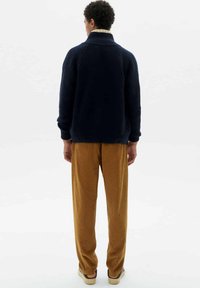 Marineblauer Strickpullover mit hohem Kragen, kombiniert mit braunen, gerippten Cordhose. Die Schuhwahl besteht aus lässigen Slippern; insgesamt ein minimalistisches Design.