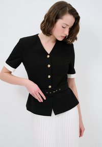 Schwarzes kurzärmeliges Oberteil mit V-Ausschnitt und goldenen Knöpfen, mit tailliertem Schnitt und einem Gürtel. Weiße Manschetten und ein Peplum-Saum.