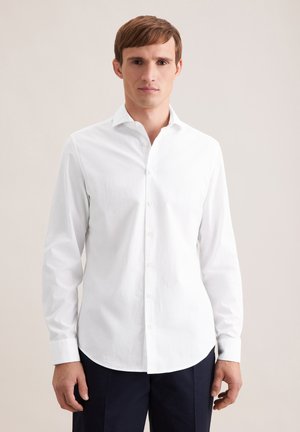 Młody mężczyzna w białej, długorękawowej koszuli na guziki i ciemnych spodniach, stojący na tle jednolitego jasnego tła.