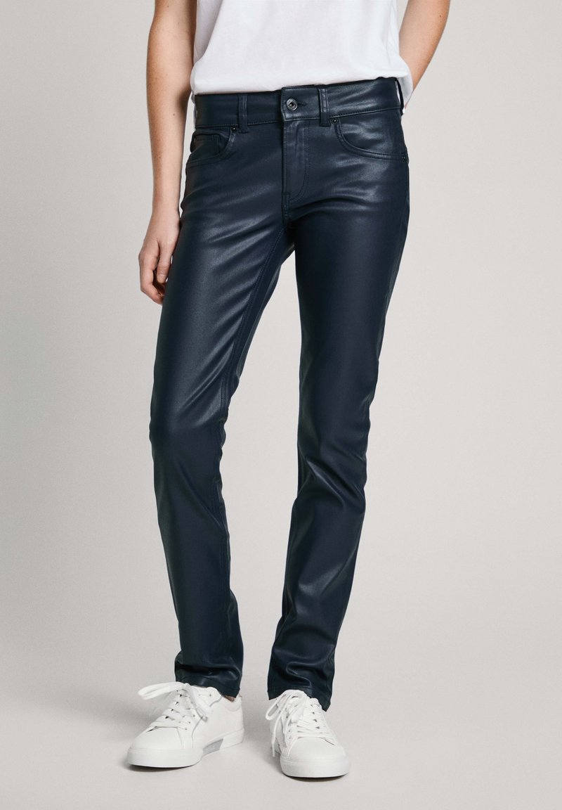 Personne portant un pantalon en cuir noir slim avec poches avant et des baskets blanches, debout devant un fond clair uni.