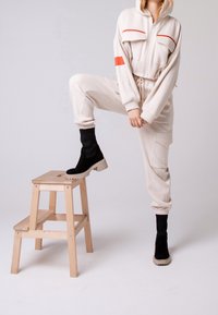 Beżowy kombinezon z polaru z kontrastowymi czerwonymi paskami, wyposażony w kaptur, zamek z przodu i kieszenie cargo. Model ma na sobie czarne skarpetki z dzianiny i buty.