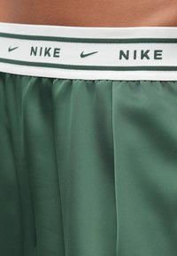 Πράσινα σορτς με λευκή μέση που φέρει επαναλαμβανόμενα σκοτεινά πράσινα λογότυπα Nike και σύμβολα swoosh στη μέση ενός ατόμου.