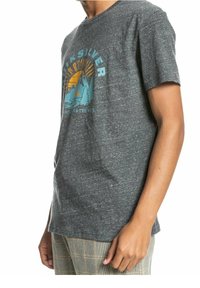 Persona vistiendo una camiseta gris oscuro de Quiksilver con un gráfico de montaña, árbol y sol, y pantalones de cuadros, en una pose relajada.