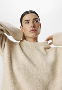 Pull en tricot beige clair, avec une coupe ample, des manches larges et une finition texturée. L'encolure est côtelée et haute, ce qui renforce l'apparence douillette.