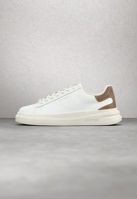 Baskets basses blanches avec patch beige au talon, design à lacets et semelle épaisse embossée du logo "GUESS" sur le côté.