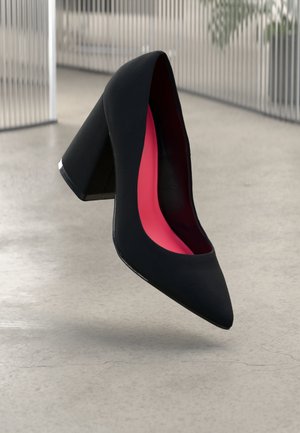 Schwarzer High-Heel-Schuh mit spitz zulaufender Zehe und klobigem Absatz, der eine leuchtend pinke Innensohle aufweist und über einem glatten, hellfarbigen Boden schwebt.