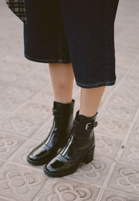 Czarne lakierowane botki na kostkę z zamkiem błyskawicznym, okrągłym noskiem i grubym obcasem. Posiadają regulowany pasek z klamrą w okolicy kostki.