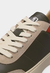 Gros plan d'une basket avec du daim beige, des panneaux en cuir vert foncé, des lacets crème et un détail rouge près du talon.