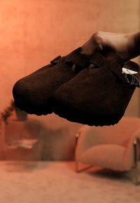 Χέρι κρατά καφέ σουέντ σανδάλια τύπου κλογκ με ιμάντες και αγκράφες, μπροστά σε υποτονικό πορτοκαλί φόντο δωματίου με καρέκλα και γλαστράκι.