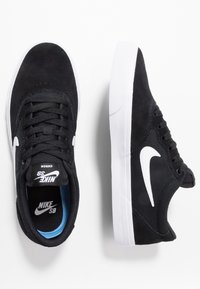 Černé semišové skate boty s bílými akcenty, s plochým šněrovacím designem a gumovou podrážkou. Uvnitř mají modrou podšívku a branding.