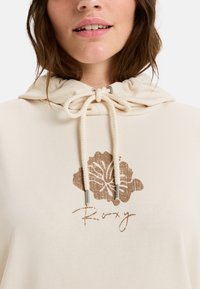 Sudadera beige con capucha ajustable, presenta un gráfico floral texturizado en marrón y la palabra "Roxy" en una fuente cursiva. Textura de tela suave.