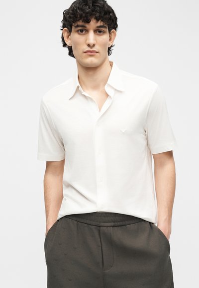 Νεαρός με σγουρά μαλλιά, φορώντας ένα λευκό πουκάμισο με κοντά μανίκια και σκούρο παντελόνι, στέκεται με τα χέρια στις τσέπες μπροστά σε απλό φόντο.