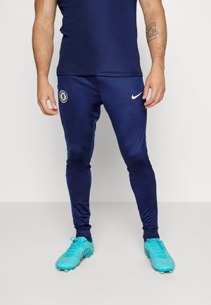 Pantaloni sportivi blu navy con un design aderente, dotati di un logo Nike bianco e un emblema del Chelsea FC. Indossati con scarpe da calcio in un vivace colore turchese.