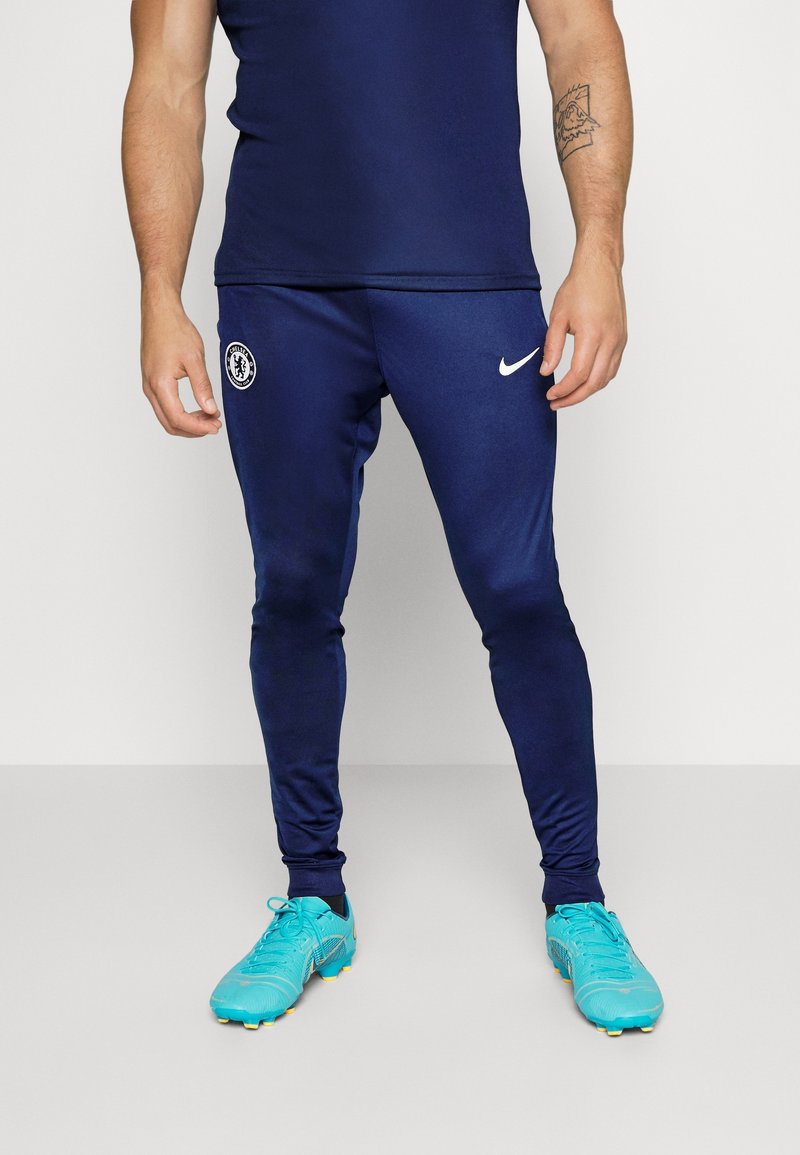 Pantalons de sport navy avec une coupe ajustée, présentant un logo Nike blanc et un emblème du Chelsea FC. Portés avec des crampons teal vif.