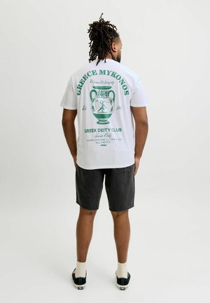 Férfi, dreadlockkal, fehér pólót visel, melyen zöld görög kerámia grafika és szöveg van, fekete rövidnadrágot, fehér zoknit és fekete sportcipőt visel, áll.