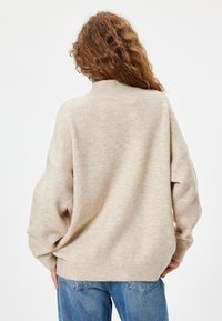 Pull en tricot beige avec un col montant et une coupe oversize. Il présente des épaules tombantes et des poignets côtelés, associé à un jean bleu.