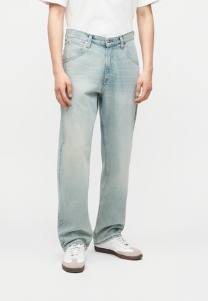 Osoba nosi svetlomodré džínsové nohavice s rovným strihom, biele tričko a biele tenisky s hnedou podrážkou, stojí na bielom pozadí.