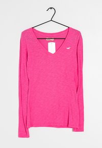 Langärmliges, V-Ausschnitt-Shirt in leuchtendem Pink, das eine weiche Textur aufweist und ein kleines weißes Logo auf der linken Brust hat. Hängt an einem schwarzen Haken.