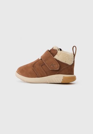 Braunes Wildleder-Kinderschuh mit einem flauschigen, hellbraunen Innenfutter, verstellbarem Klettverschluss und flexibler Gummisohle. Abgerundete Zehenpartie und leichter Absatz.